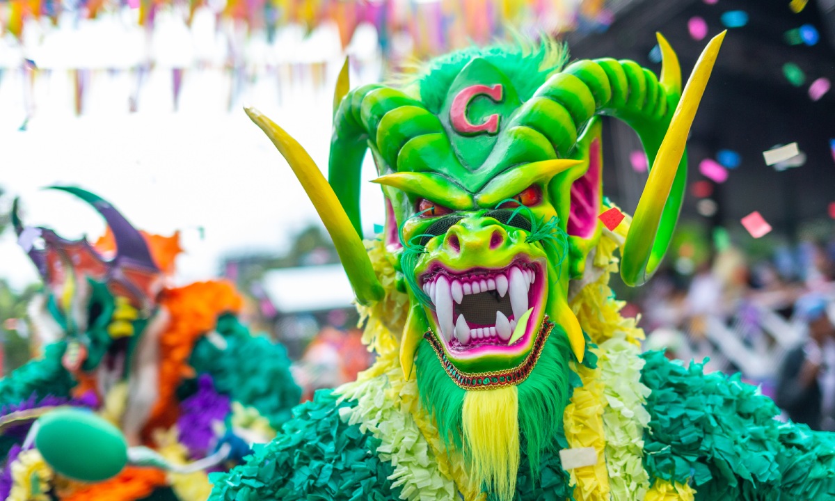 Primer plano de un personaje del carnaval dominicano con una máscara verde neón de expresión feroz, grandes cuernos amarillos y una "G" en la frente. El traje está hecho de capas de papel o tela verde y amarilla, y el fondo muestra confeti cayendo y otras máscaras difuminadas.