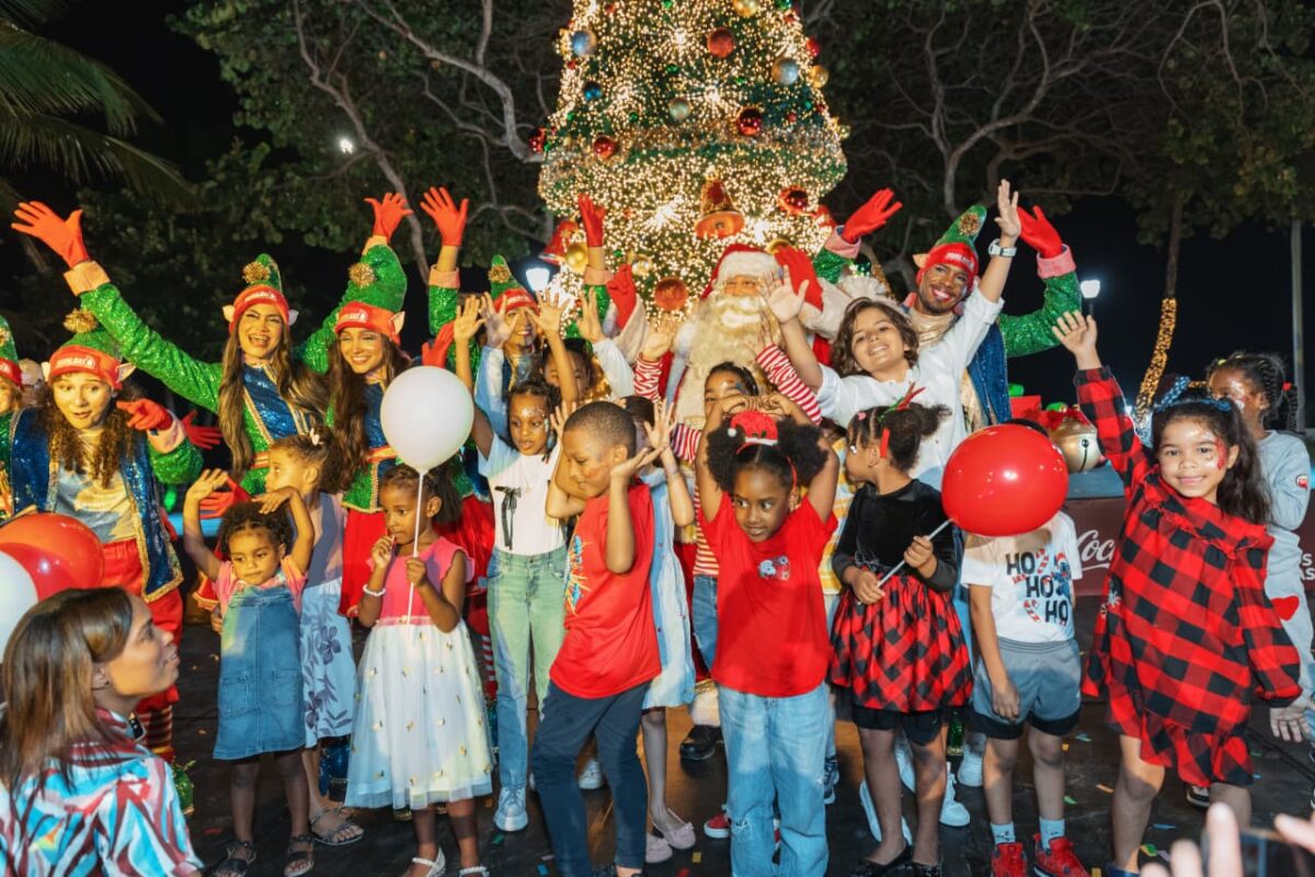Fotografía nocturna en un parque decorado. Un gran grupo de niños y niñas posa sonriente frente a un árbol de Navidad gigante y muy iluminado. En el centro está Santa Claus, rodeado de personajes disfrazados de elfos verdes y asistentes. Los niños sostienen globos rojos y blancos, y muchos levantan las manos con alegría.