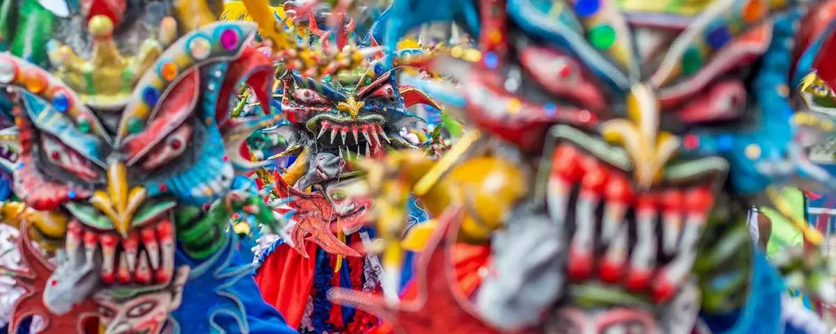 Primer plano vibrante de varios Diablos Cojuelos dominicanos luciendo máscaras coloridas de rasgos exagerados, dientes afilados y cuernos, decoradas con lentejuelas y plumas durante el Desfile Nacional de Carnaval.