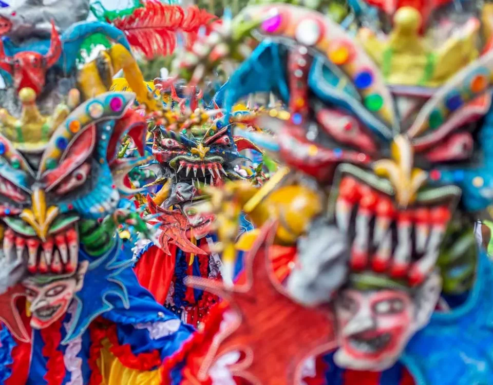 Primer plano vibrante de varios Diablos Cojuelos dominicanos luciendo máscaras coloridas de rasgos exagerados, dientes afilados y cuernos, decoradas con lentejuelas y plumas durante el Desfile Nacional de Carnaval.